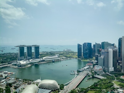 Aerial view of a modern urban landscape with a prominent hotel featuring three towers connected by an elevated platform. Surrounding the hotel are sleek skyscrapers and a body of water housing several small boats. The scene includes lush greenery and a large ferris wheel in the distance.