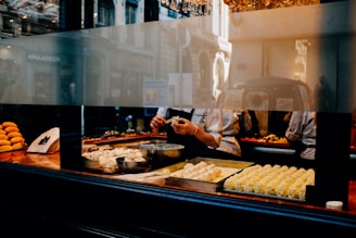 A bakery kitchen is visible through a large window, where several chefs are at work. One chef is preparing pastries or bread on a wooden countertop lined with various baked goods. Several trays with dough and baked items are displayed prominently in the foreground. The setting appears to be a busy urban street, reflecting city buildings on the glass.