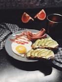 A plate of breakfast food including a sunny side up egg with hot sauce drizzled on top, sliced avocado on toast with seasoning, and slices of sausage. There are two halves of a grapefruit in the background along with a cup on a dark surface, and a checkered cloth nearby.