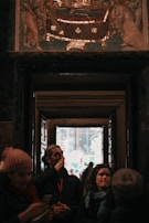 A group of people standing inside a darkened interior space, possibly a museum or historical site. They seem to be observing an artifact or artwork displayed above a doorway. The artwork appears to be a mosaic or painting with religious or historical themes. Light filters through an open door in the background, creating a strong contrast between the dark indoor space and the bright exterior.