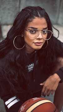 A person with long dark hair and large hoop earrings is wearing eyeglasses. They are dressed in a black outfit and are holding a basketball.