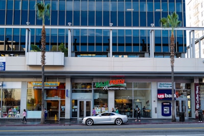 A street view of a modern building with reflective glass windows. The ground level features various businesses including a pretzel shop, a Mexican grill, and a bank. A silver sports car is parked on the street, and several people with small bags are walking along the sidewalk. There are a few palm trees lining the street.