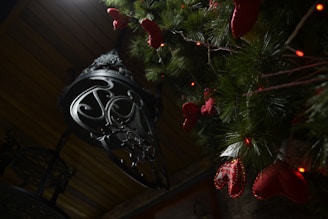 A darkly lit scene featuring a decorative metal lantern with intricate designs hanging from a wooden ceiling. Next to the lantern, a green pine tree branch is adorned with red ornamental hearts and small red lights, suggesting a festive decoration.