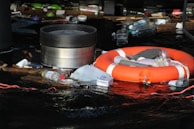 A collection of debris and garbage, including plastic bottles and containers, is floating on dark water. Among the waste, there is a bright orange life preserver. The area appears cluttered with various types of plastic waste, highlighting environmental pollution.