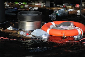 A collection of debris and garbage, including plastic bottles and containers, is floating on dark water. Among the waste, there is a bright orange life preserver. The area appears cluttered with various types of plastic waste, highlighting environmental pollution.