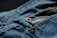Close-up view of blue denim fabric with a focus on a pocket that features a red logo tag. Copper rivets are visible, highlighting the stitching and texture of the material.