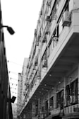 A black and white photograph of an urban street scene featuring a large, multi-story building with numerous air conditioning units attached to the exterior. The building is seen from an angled perspective, emphasizing its tall structure. There are string lights hanging across the street and Korean signage on the lower levels.