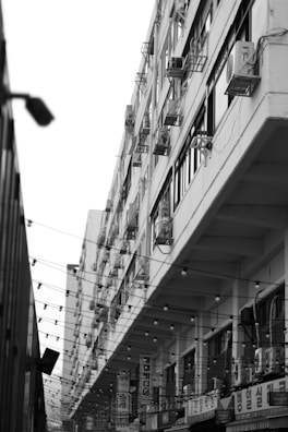 A black and white photograph of an urban street scene featuring a large, multi-story building with numerous air conditioning units attached to the exterior. The building is seen from an angled perspective, emphasizing its tall structure. There are string lights hanging across the street and Korean signage on the lower levels.
