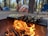 A campsite in a forested area with several tents visible among tall trees. A grill over an open fire contains corn on the cob wrapped in husks and a tray with skewered vegetables and meats. The flames are bright and prominent, casting a warm glow. Camping chairs are also seen nearby.
