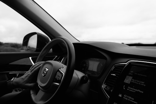 A driver's hand rests on the steering wheel of a car, viewed from the perspective of someone sitting in the driver's seat. The dashboard and part of the radio display are visible, showing some digital information. The interior is modern with sleek lines and a view of the outside landscape in the background through the side window.