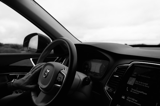 A driver's hand rests on the steering wheel of a car, viewed from the perspective of someone sitting in the driver's seat. The dashboard and part of the radio display are visible, showing some digital information. The interior is modern with sleek lines and a view of the outside landscape in the background through the side window.