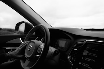 A driver's hand rests on the steering wheel of a car, viewed from the perspective of someone sitting in the driver's seat. The dashboard and part of the radio display are visible, showing some digital information. The interior is modern with sleek lines and a view of the outside landscape in the background through the side window.