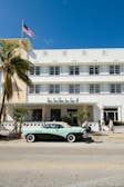 A vintage convertible car is parked on a street in front of a white Art Deco-style hotel. The building has a geometric design with horizontal lines and windows. A palm tree is on the left side and the American flag is waving on top of the building. A few pedestrians walk along the sidewalk, enjoying the sunny day.