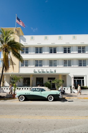 A vintage convertible car is parked on a street in front of a white Art Deco-style hotel. The building has a geometric design with horizontal lines and windows. A palm tree is on the left side and the American flag is waving on top of the building. A few pedestrians walk along the sidewalk, enjoying the sunny day.