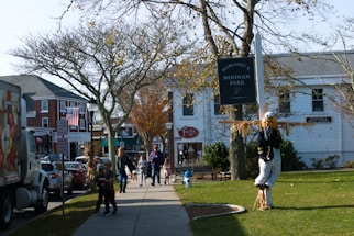 A street scene features a park sign reading 'Noonan Park' with a scarecrow displayed next to it. People, including adults and children, walk along the sidewalk lined with bare trees. Buildings with storefronts and an American flag are visible in the background on a sunny day.