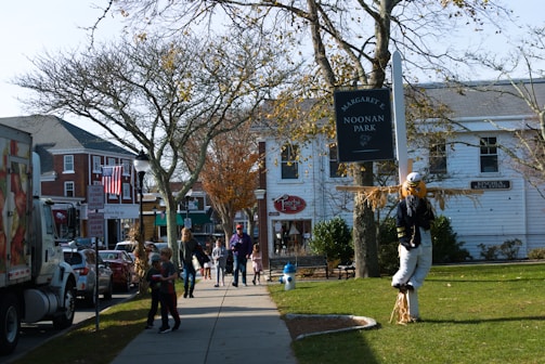 A street scene features a park sign reading 'Noonan Park' with a scarecrow displayed next to it. People, including adults and children, walk along the sidewalk lined with bare trees. Buildings with storefronts and an American flag are visible in the background on a sunny day.