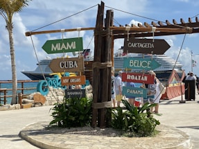 A collection of colorful directional signs pointing towards various destinations, including Miami, Cuba, and Panama, is prominently displayed. Behind the signs, a large cruise ship is docked at a port filled with people walking around under a clear blue sky.
