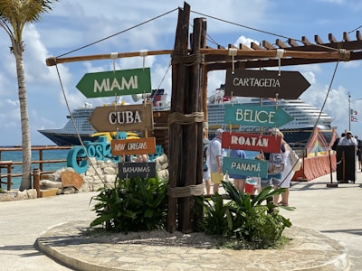 A collection of colorful directional signs pointing towards various destinations, including Miami, Cuba, and Panama, is prominently displayed. Behind the signs, a large cruise ship is docked at a port filled with people walking around under a clear blue sky.