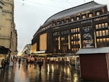 A bustling urban street scene in front of a large department store adorned with bright, festive lights. Several people walk along the wet, reflective pavement, and small market stalls are set up to the right. The sky is overcast, suggesting recent rain or ongoing drizzle.