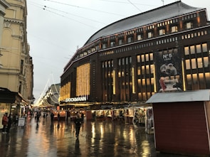 A bustling urban street scene in front of a large department store adorned with bright, festive lights. Several people walk along the wet, reflective pavement, and small market stalls are set up to the right. The sky is overcast, suggesting recent rain or ongoing drizzle.
