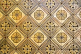 A geometric pattern of vintage floor tiles featuring floral motifs in earthy tones. The tiles are arranged in a repetitive diamond layout, with intricate flower designs at the center of each tile. The aged appearance of the tiles suggests historical or classical architecture.