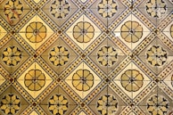 A geometric pattern of vintage floor tiles featuring floral motifs in earthy tones. The tiles are arranged in a repetitive diamond layout, with intricate flower designs at the center of each tile. The aged appearance of the tiles suggests historical or classical architecture.