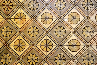 A geometric pattern of vintage floor tiles featuring floral motifs in earthy tones. The tiles are arranged in a repetitive diamond layout, with intricate flower designs at the center of each tile. The aged appearance of the tiles suggests historical or classical architecture.