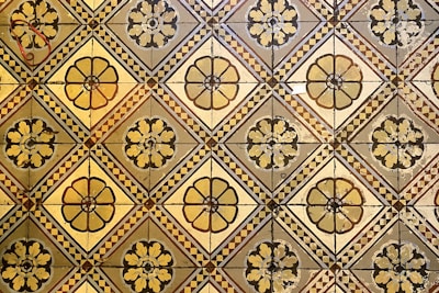 A geometric pattern of vintage floor tiles featuring floral motifs in earthy tones. The tiles are arranged in a repetitive diamond layout, with intricate flower designs at the center of each tile. The aged appearance of the tiles suggests historical or classical architecture.