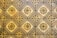 A geometric pattern of vintage floor tiles featuring floral motifs in earthy tones. The tiles are arranged in a repetitive diamond layout, with intricate flower designs at the center of each tile. The aged appearance of the tiles suggests historical or classical architecture.