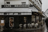A quaint Parisian cafe called 'Le Saint Jean' with a vintage aesthetic. The exterior features striped awnings and a classic sign that reads 'Cafe Brasserie'. There are rattan chairs and small tables on the sidewalk, suggesting outdoor seating. The window displays advertise services such as continuous dining and jazz concerts. The street is wet, indicating recent rain, and a couple of people are seen walking by.