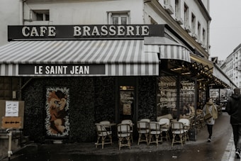 A quaint Parisian cafe called 'Le Saint Jean' with a vintage aesthetic. The exterior features striped awnings and a classic sign that reads 'Cafe Brasserie'. There are rattan chairs and small tables on the sidewalk, suggesting outdoor seating. The window displays advertise services such as continuous dining and jazz concerts. The street is wet, indicating recent rain, and a couple of people are seen walking by.