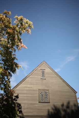 A wooden house with rustic, lattice-style windows stands against a clear blue sky. To the left, green foliage from a nearby tree partly frames the view, while the shadows of the tree fall across the side of the building.