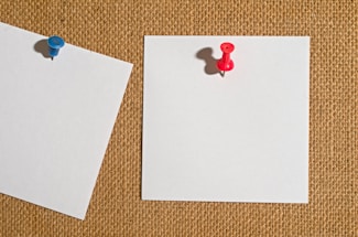 Two blank white notes pinned to a burlap surface, one with a blue pushpin and the other with a red pushpin.