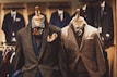 Two mannequins are dressed in elegant men's suits. The left mannequin sports a layered outfit with a brown textured jacket, blue waistcoat, and patterned tie, accented by a vibrant pocket square. The right mannequin features a checkered gray suit with a matching tie, showcasing a classic and sophisticated look. Various other suits are visible in the softly focused background of the clothing store.