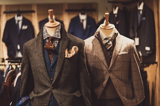 Two mannequins are dressed in elegant men's suits. The left mannequin sports a layered outfit with a brown textured jacket, blue waistcoat, and patterned tie, accented by a vibrant pocket square. The right mannequin features a checkered gray suit with a matching tie, showcasing a classic and sophisticated look. Various other suits are visible in the softly focused background of the clothing store.