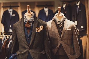 Two mannequins are dressed in elegant men's suits. The left mannequin sports a layered outfit with a brown textured jacket, blue waistcoat, and patterned tie, accented by a vibrant pocket square. The right mannequin features a checkered gray suit with a matching tie, showcasing a classic and sophisticated look. Various other suits are visible in the softly focused background of the clothing store.