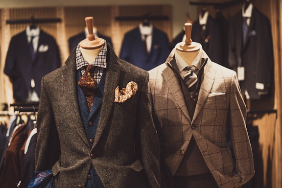 Two mannequins are dressed in elegant men's suits. The left mannequin sports a layered outfit with a brown textured jacket, blue waistcoat, and patterned tie, accented by a vibrant pocket square. The right mannequin features a checkered gray suit with a matching tie, showcasing a classic and sophisticated look. Various other suits are visible in the softly focused background of the clothing store.