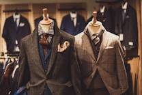 Two mannequins are dressed in elegant men's suits. The left mannequin sports a layered outfit with a brown textured jacket, blue waistcoat, and patterned tie, accented by a vibrant pocket square. The right mannequin features a checkered gray suit with a matching tie, showcasing a classic and sophisticated look. Various other suits are visible in the softly focused background of the clothing store.