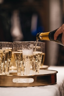 A hand pours champagne from a bottle into tall, elegant glasses arranged on a tray. The bubbly liquid is frothy and clear, creating a festive atmosphere.
