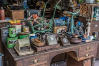 A cluttered antique shop display features a variety of vintage items on a wooden desk, including rotary dial telephones, an abacus, and old tin cans with colorful labels. A whimsical toy figure sits amidst the items, alongside a small blue tricycle and various decorative objects. The setting suggests a collection of retro and nostalgic items, with rich textures and an eclectic mix of colors.