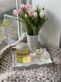 A cozy scene features a glass teapot filled with light yellow tea and a matching teacup on a decorative tray. Next to the tea set is a white vase filled with pink and white tulips. The arrangement is placed on a chunky knitted blanket by a window, creating a comforting and serene atmosphere.