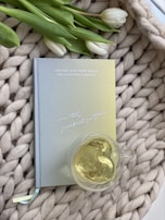 A textured, woven surface with a pastel-colored book titled 'Explore Your Inner World for Happiness & Growth' is accompanied by a glass cup filled with light-colored tea. A few white tulips with green leaves provide an elegant touch.