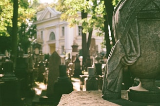 A cemetery scene with numerous gravestones and monuments casting shadows under the sunlight. In the foreground, an ornate stone sculpture appears with draped fabric details. A white, classical-style building with columns and an arched entrance stands in the background, partially obscured by trees.