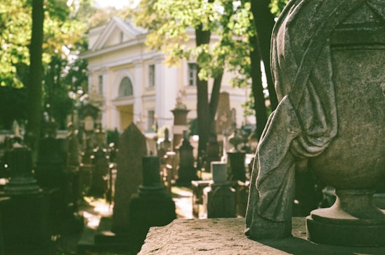 A cemetery scene with numerous gravestones and monuments casting shadows under the sunlight. In the foreground, an ornate stone sculpture appears with draped fabric details. A white, classical-style building with columns and an arched entrance stands in the background, partially obscured by trees.