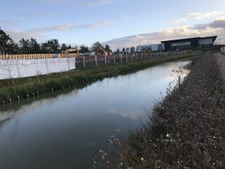 A calm water canal runs parallel to a construction site with cranes and building materials. The area is enclosed by a metal fence with white fabric banners. In the background, a large industrial building is visible. Green vegetation lines the canal, and the sky is partly cloudy, suggesting early morning or late afternoon light.