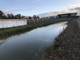 A calm water canal runs parallel to a construction site with cranes and building materials. The area is enclosed by a metal fence with white fabric banners. In the background, a large industrial building is visible. Green vegetation lines the canal, and the sky is partly cloudy, suggesting early morning or late afternoon light.
