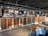 A spacious and modern bar area with a long wooden counter, featuring several drink taps and a coffee machine. The back wall is adorned with black and white photographs, adding a vintage touch. Blue and wooden chairs are set around small round tables in the seating area. Overhead lighting provides a warm and inviting atmosphere.