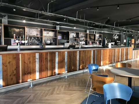 A spacious and modern bar area with a long wooden counter, featuring several drink taps and a coffee machine. The back wall is adorned with black and white photographs, adding a vintage touch. Blue and wooden chairs are set around small round tables in the seating area. Overhead lighting provides a warm and inviting atmosphere.