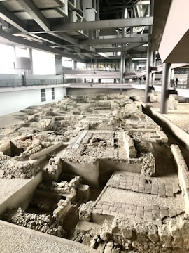 An archaeology site featuring ancient ruins with stone and brick masonry is located indoors within a modern architectural structure. The area is lit by natural and artificial light, highlighting the complexity and depth of the uncovered remnants. Elevated walkways are visible in the background, allowing visitors to view the site from above.