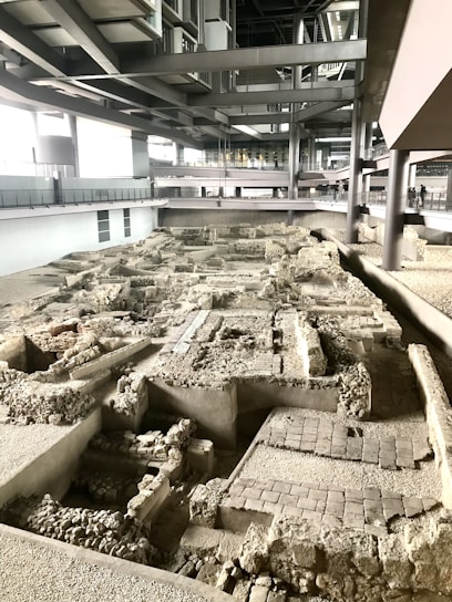 An archaeology site featuring ancient ruins with stone and brick masonry is located indoors within a modern architectural structure. The area is lit by natural and artificial light, highlighting the complexity and depth of the uncovered remnants. Elevated walkways are visible in the background, allowing visitors to view the site from above.